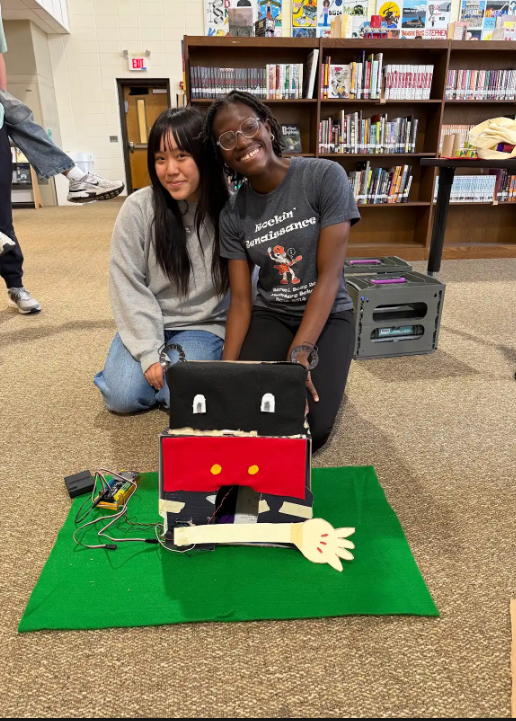 Two individuals crouch next to a homemade robot, positioned on a green mat in a library.