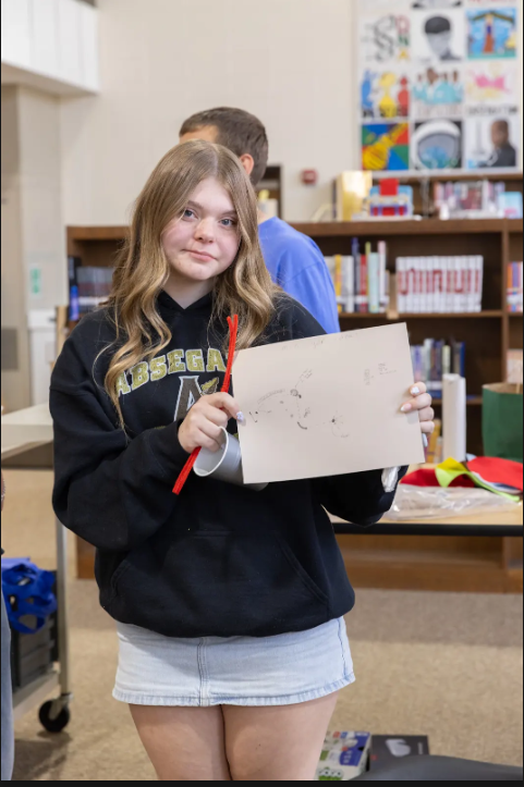 Girl holding a piece of paper in her hands, standing in a library. A man stands behind her, near a bookshelf.