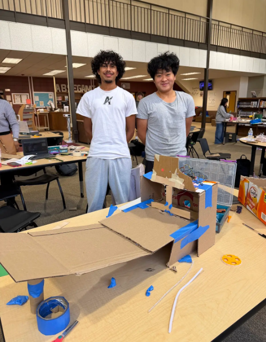 Two individuals in a library stand behind a table displaying a cardboard project. Other tables and chairs are visible in the background.
