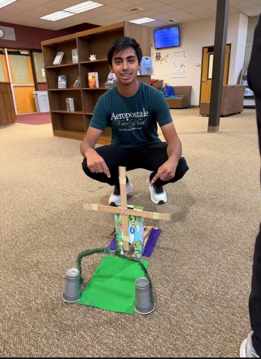 A man crouches with a homemade wooden cart, holding a small box, in a room with bookshelves.