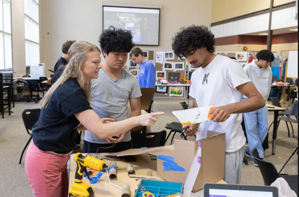 A group of people in a room with tools and supplies, a woman points at a piece of paper.