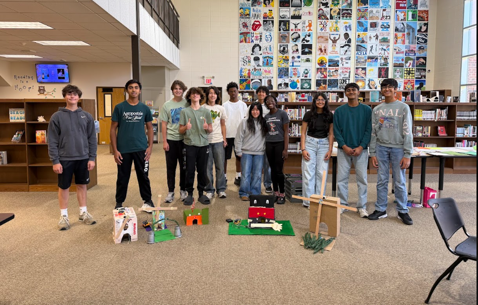 Several students pose with various models on a carpeted floor, surrounded by library shelves, a monitor, and chairs.