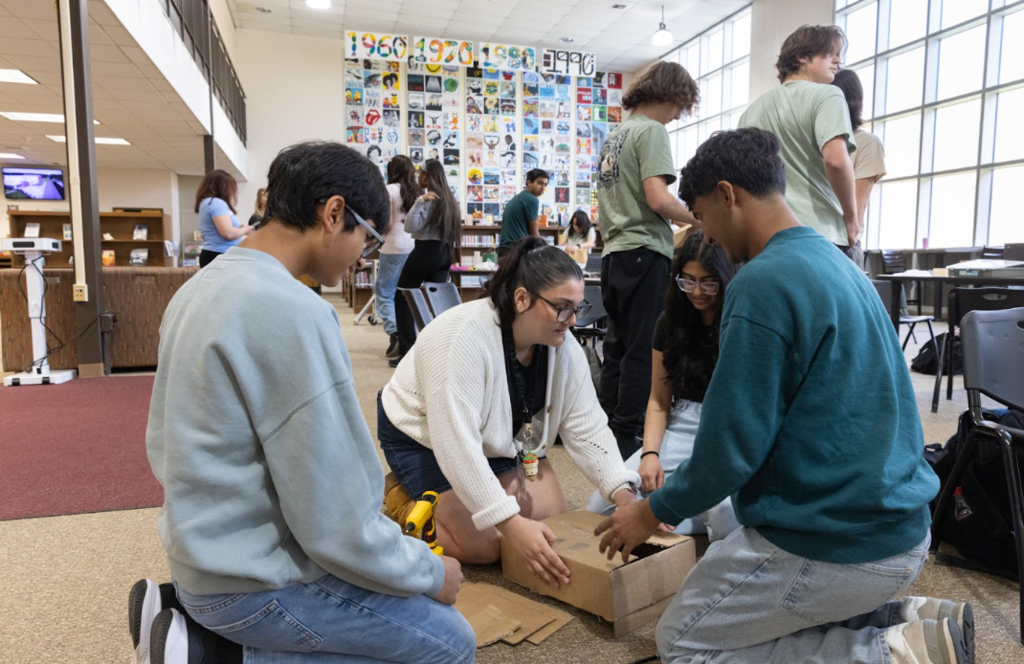 A group of students in a classroom, working together on a project with cardboard. Background features a colorful wall display.