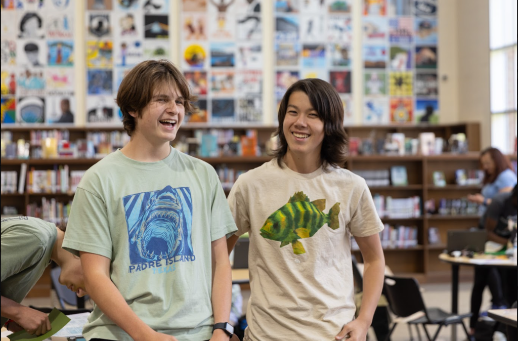 Two boys stand in a library, smiling. One wears a shark t-shirt, the other a fish. Shelves filled with books and tables are in the background.