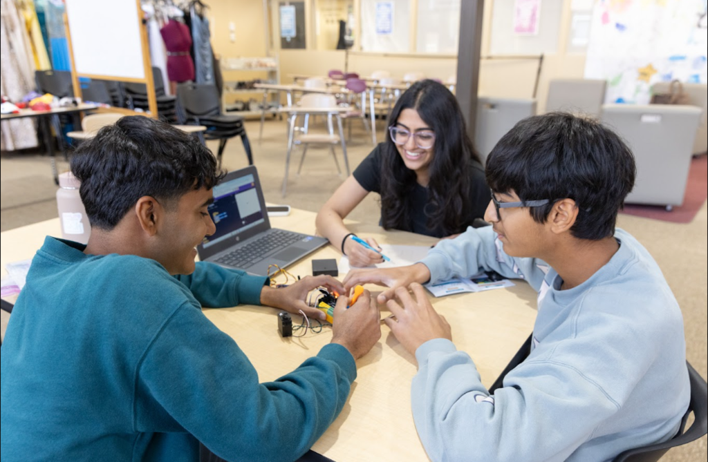 Three people at a table; two boys using electronics and a girl sitting behind them with a smile.