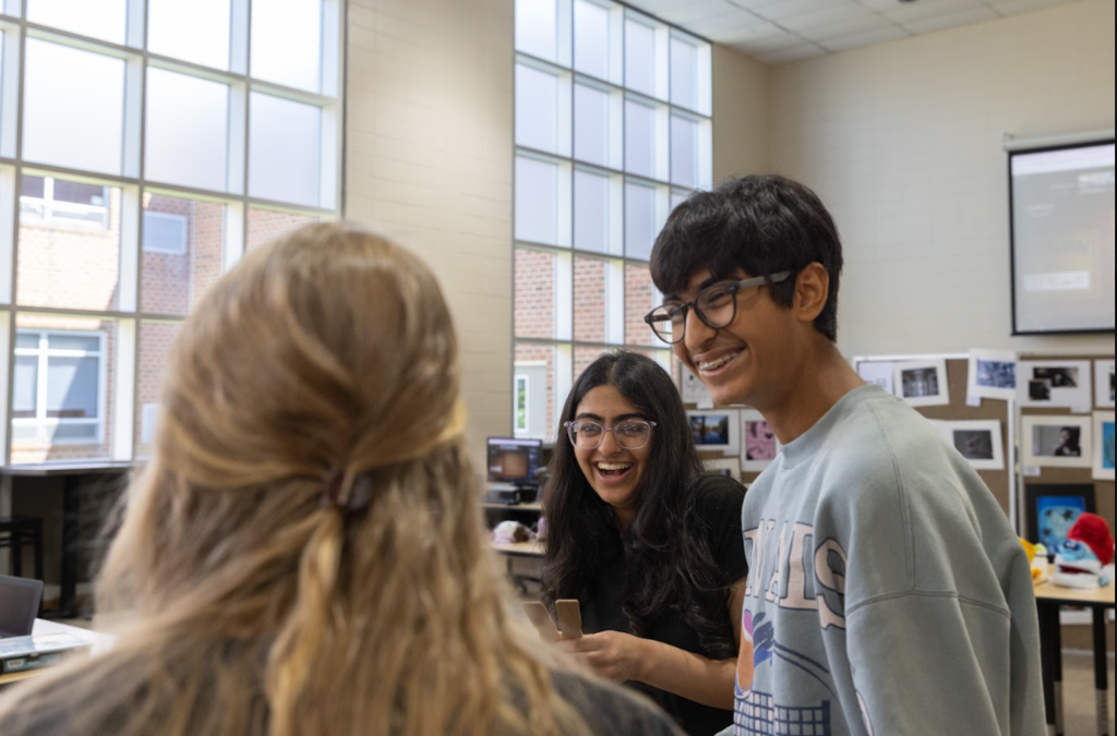 Three individuals in a room with a large window, smiling and talking. The person in the foreground has a ponytail.
