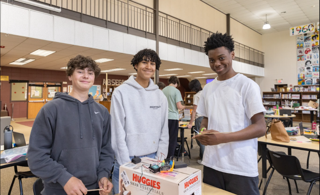 Three young men are standing next to a box in a room with tables and chairs.