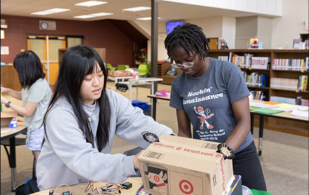 Two students in a library work on a project using a cardboard box and electronic components.