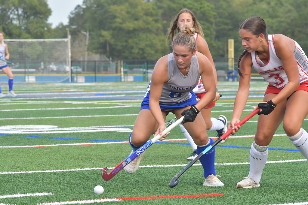 Four players wearing hockey gear on a field, two with hockey sticks, one chasing a ball.