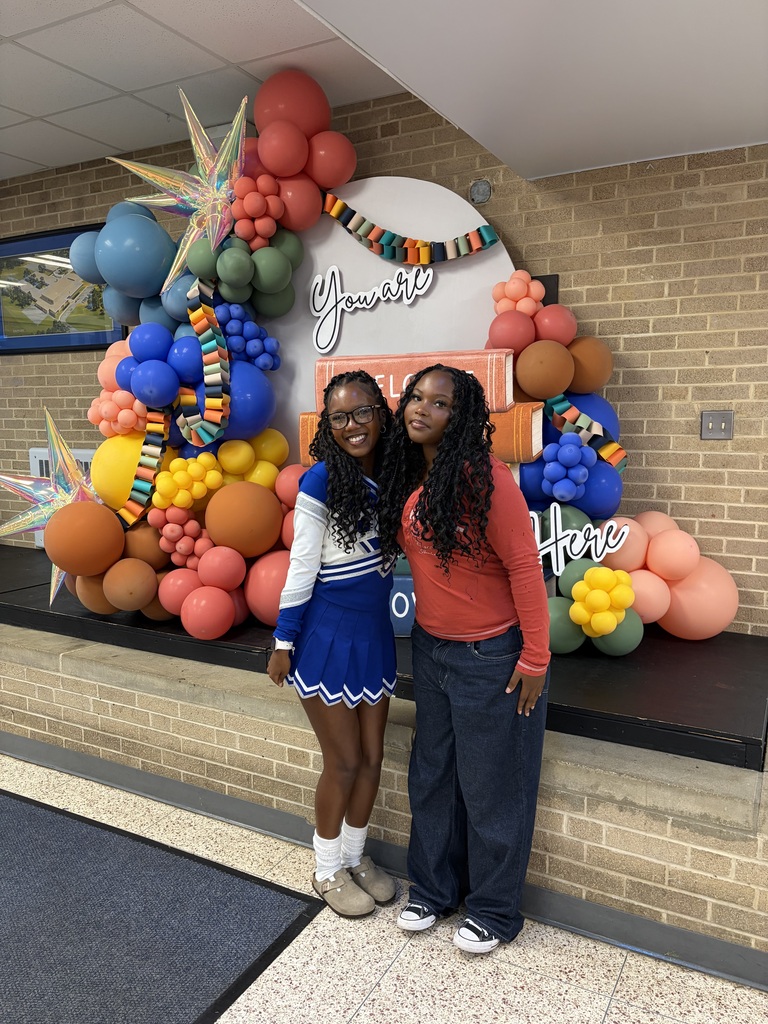 Two women, one in a cheerleading uniform, stand in front of a colorful balloon arch with text.