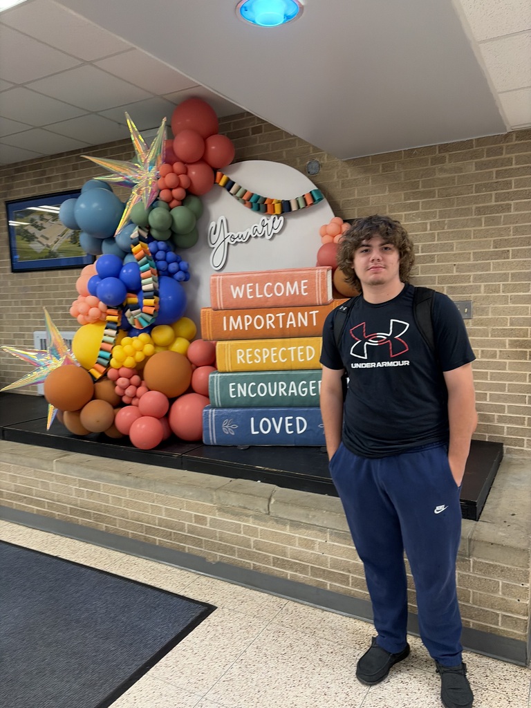 Man in black shirt and blue pants stands in front of colorful balloons and books.