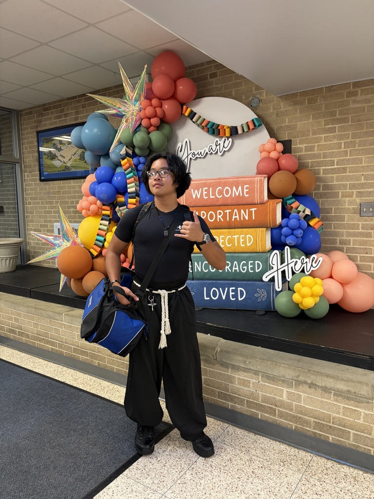 A person in black clothes poses in front of a decorative wall with colorful balloons and books.