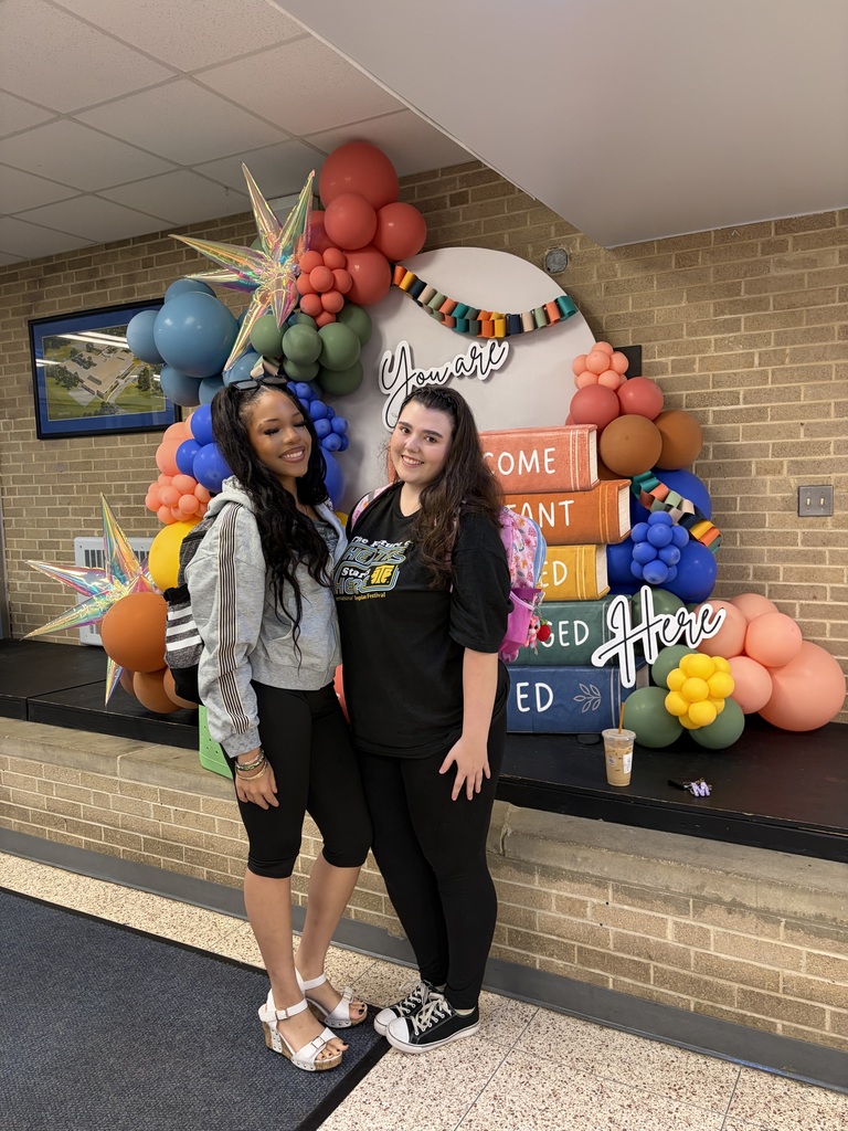 Two women stand smiling next to a colorful backdrop with balloons, text, and a framed picture.