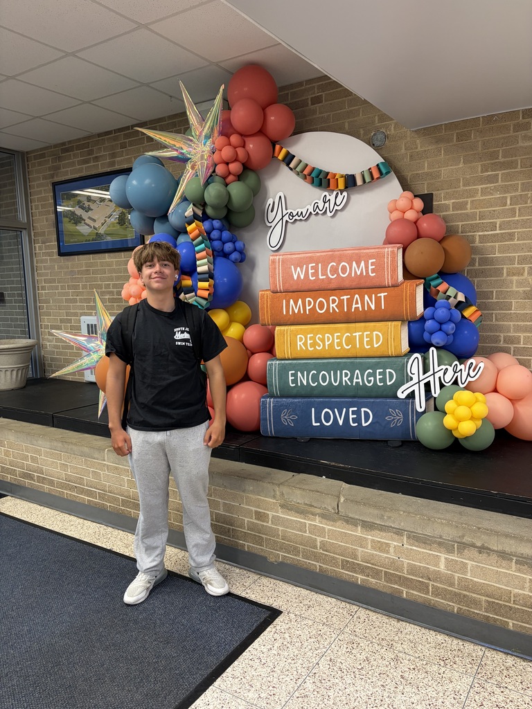 Person in black t-shirt, gray pants, white sneakers stands before a colorful balloon display. Words "Welcome, Important, Respected, Encouraged, Loved" written on books.
