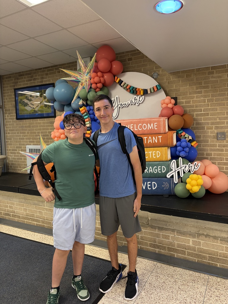 Two people stand in front of a colorful balloon display and a sign reading "Welcome". They wear casual clothing.