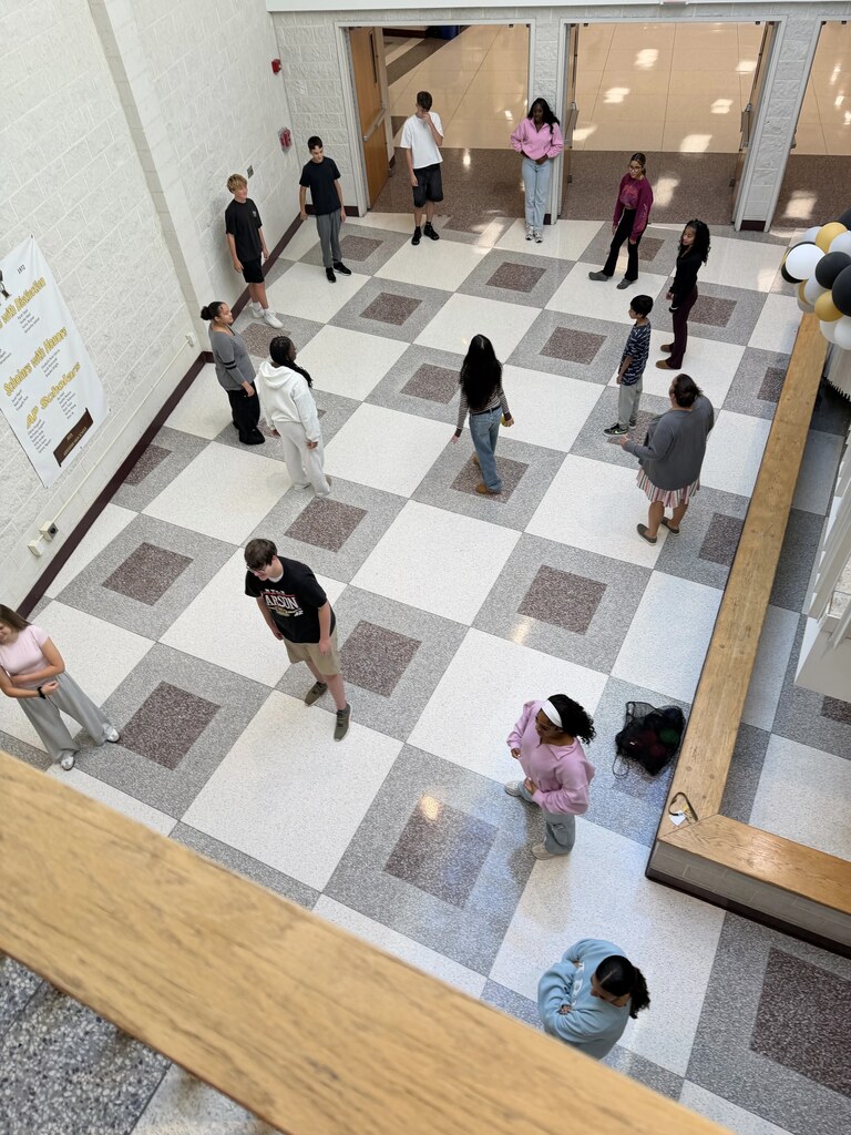 A group of people in a hall with a white and gray checkered floor, near a wall with posters.