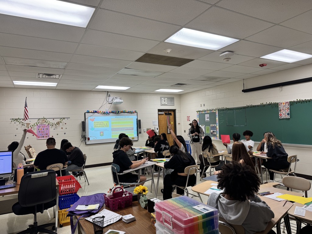 Classroom with students seated at desks, some using laptops. A chalkboard and projector screen are at the front. Tables have colorful folders, papers, and flowers.
