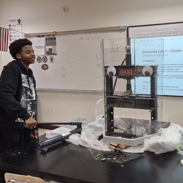 A person stands next to a table with a mechanical device in a classroom. A projector screen displays a diagram.
