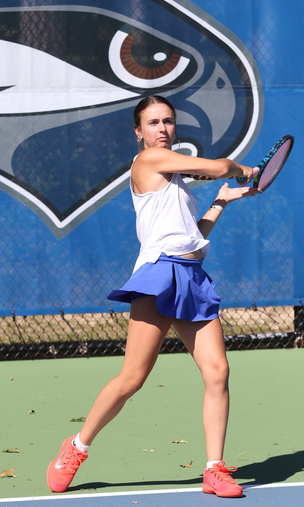 A female tennis player with a white tank top and blue skirt swings a tennis racket on a tennis court.