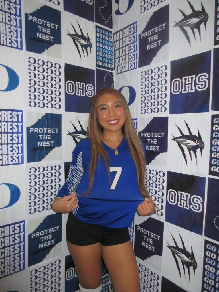 Woman with long hair wearing a blue jersey with the number 7, black shorts, and white socks, posing in front of a wall.