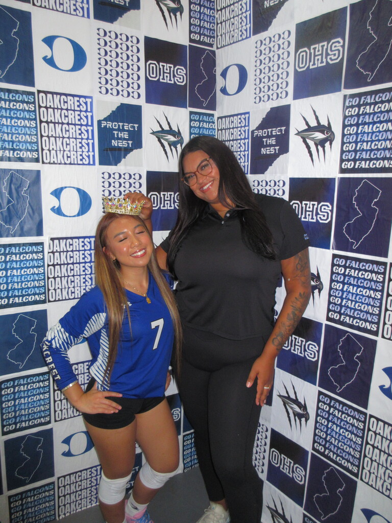 Two women, one wearing a blue jersey with the number 7, stand by a wall with blue and white designs.