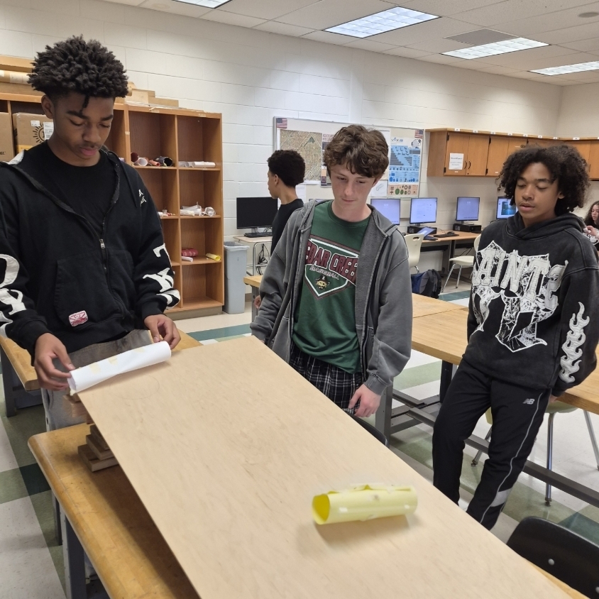 Three students work on a project at a table in a classroom with wooden shelves.