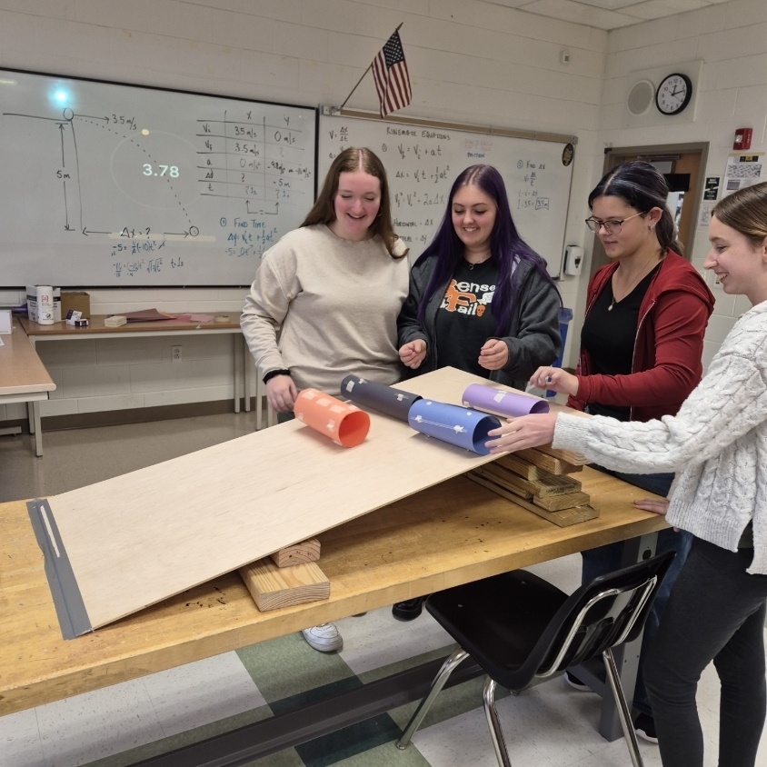 Three students collaborate on a science project in a classroom. They are working with a ramp and several objects.