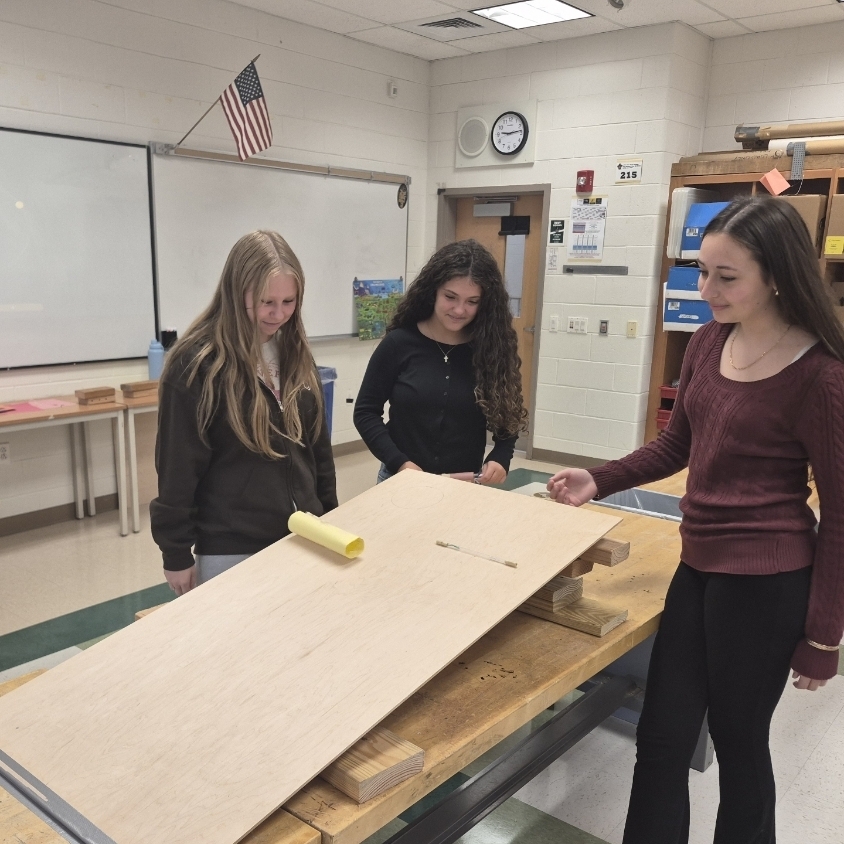 Three females in a room with a white board, a clock, and shelves with books.