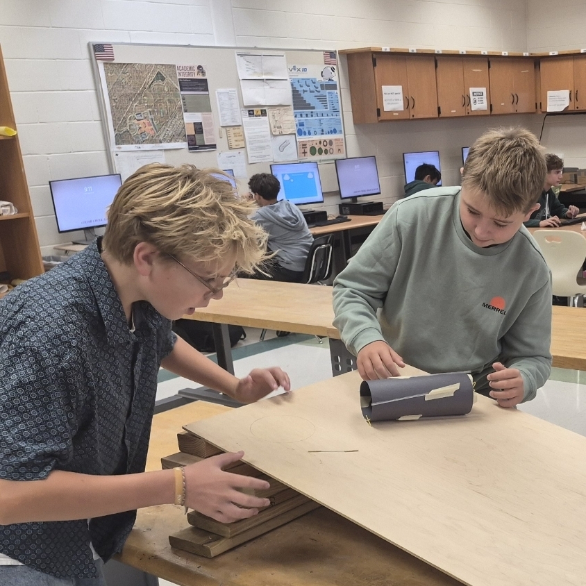 Two students working on a project in a classroom. They are surrounded by computers and tables.