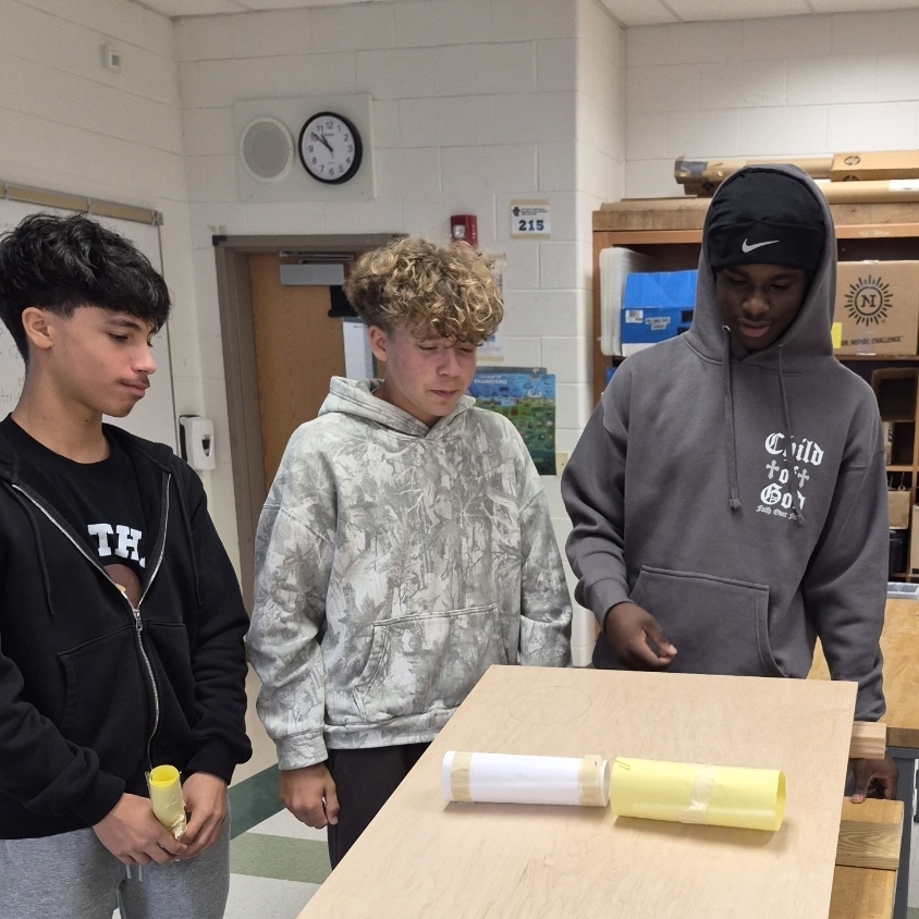 Three young males examine an object on a table in a room. One wears a black hoodie.