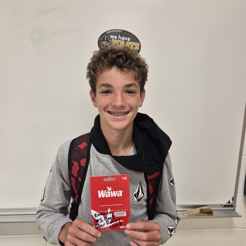 Teen wearing a gray top and a backpack holding a red card with Wawa logo in a classroom.