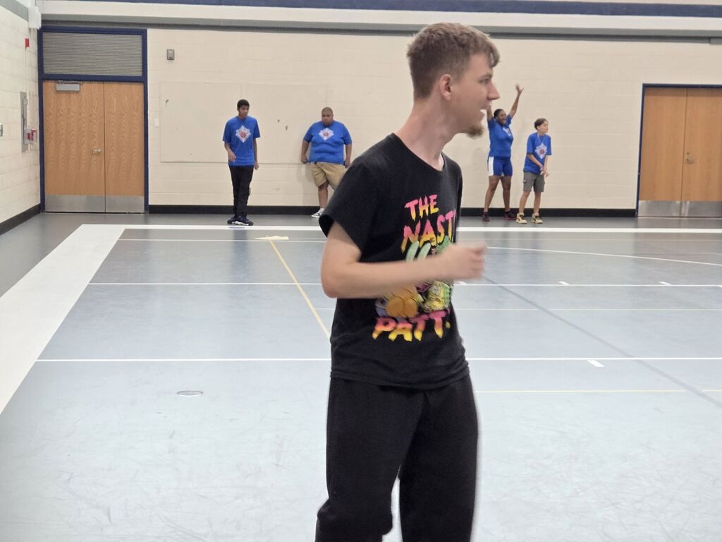 A man in a black t-shirt runs across a gym floor. Behind him, three people in blue shirts are standing.