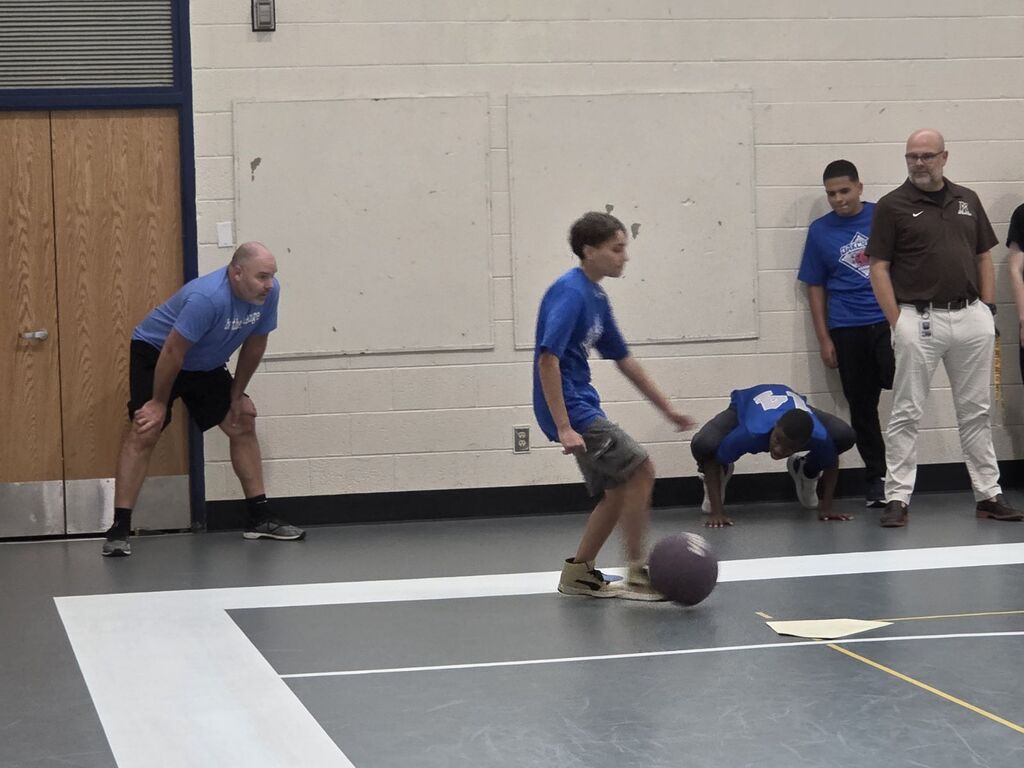 Several people on a gym floor. Two men play with a ball, while others stand behind them.