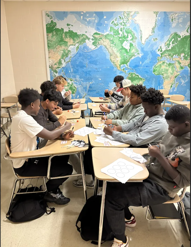 Students sit at desks, facing a large world map on the wall, working on papers. Some wear sneakers.