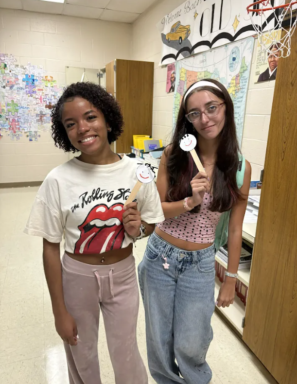 Two women pose in a hallway. One holds a guitar pick and wears a Rolling Stones shirt. The other wears a headband and holds a foam guitar.