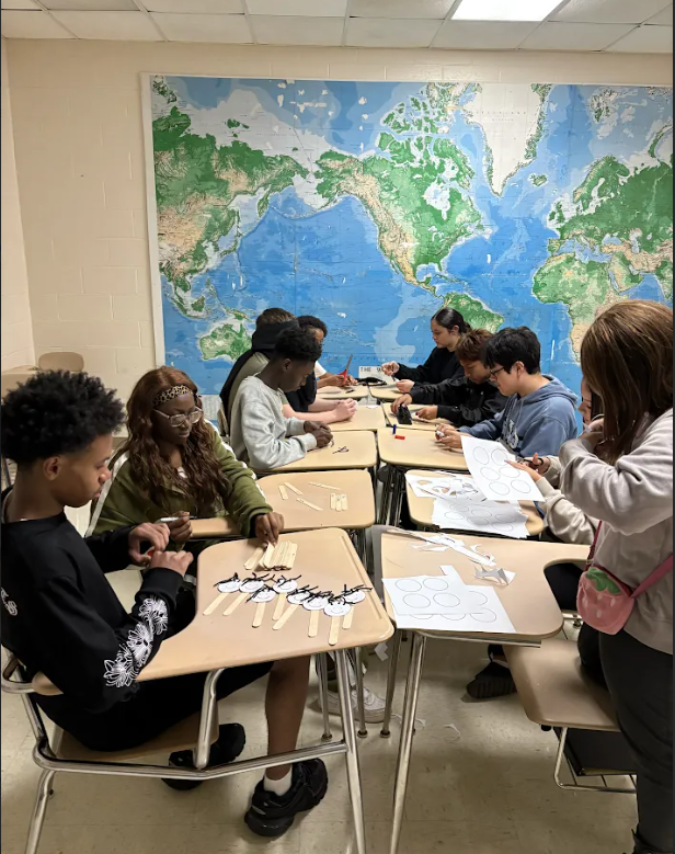 A classroom with a large world map. Students seated at desks work on projects. Papers and tools on desks.