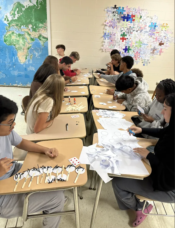 Students seated at desks in a classroom, with various art materials. A map and puzzle pieces on the wall.