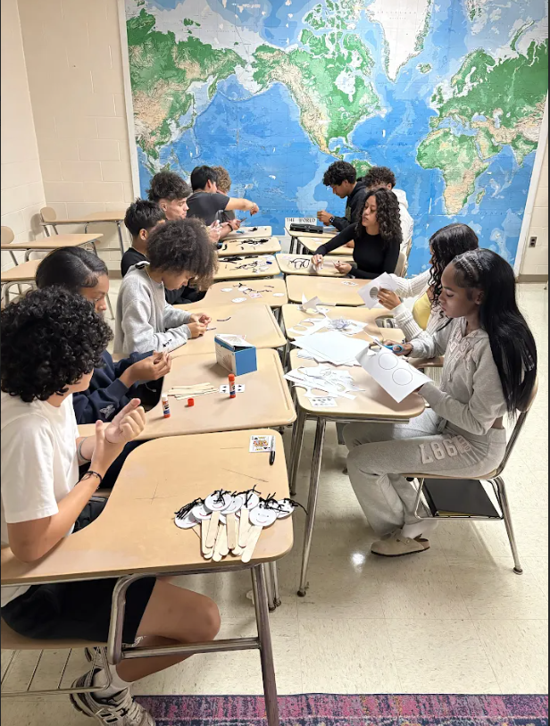 Students in a classroom with a world map on the wall, seated at desks, engaged in an activity.