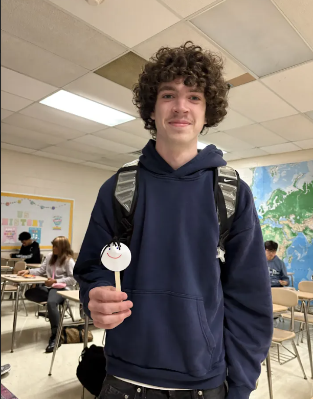 A young person in a blue hoodie stands in a classroom, holding a dessert stick with a smiley face.
