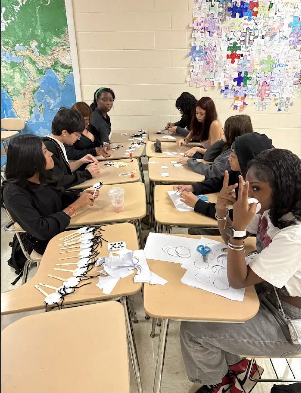 Several students seated at desks, crafting with scissors and paper in a classroom. A world map and puzzle-piece art adorn the background.
