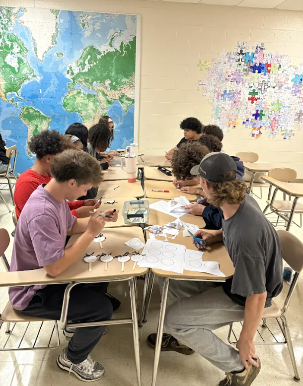 Several students work on projects at tables in a classroom, with a world map and puzzle-piece mural in the background.