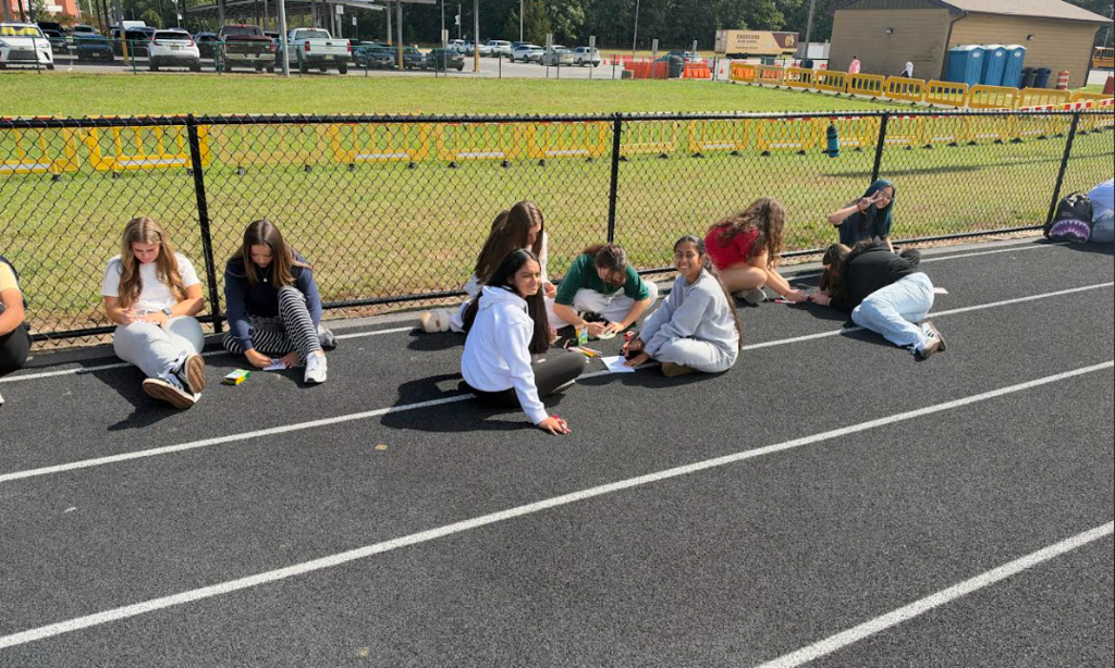 A group of people sitting on a track, some wearing white, near a fence and a grassy field.