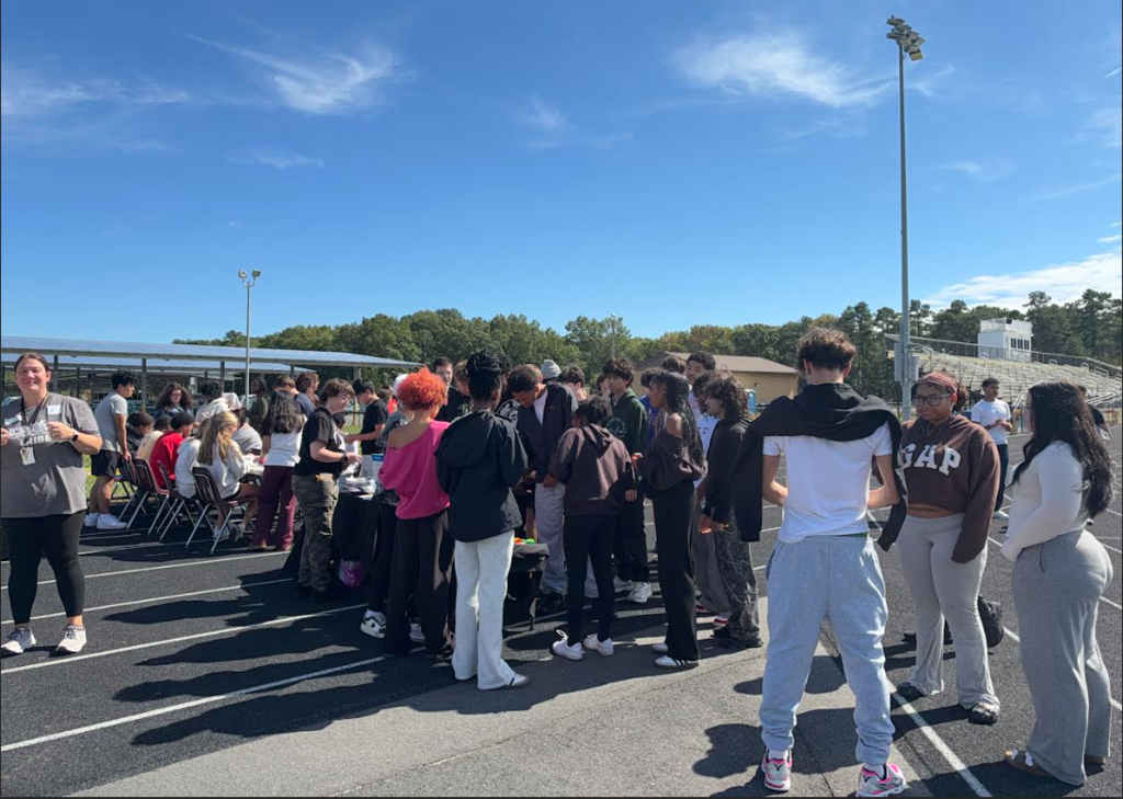 A group of individuals stands in a line on a paved surface. Some wear sneakers. Trees and a blue sky are in the background.