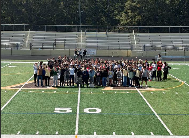 A large group of people stands on a football field, with a 50-yard line marker. Bleachers are in the background.