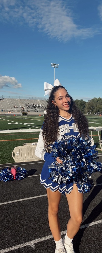 A person in a cheerleader's uniform stands on a football field holding blue and white pom-poms, with bleachers in the background.
