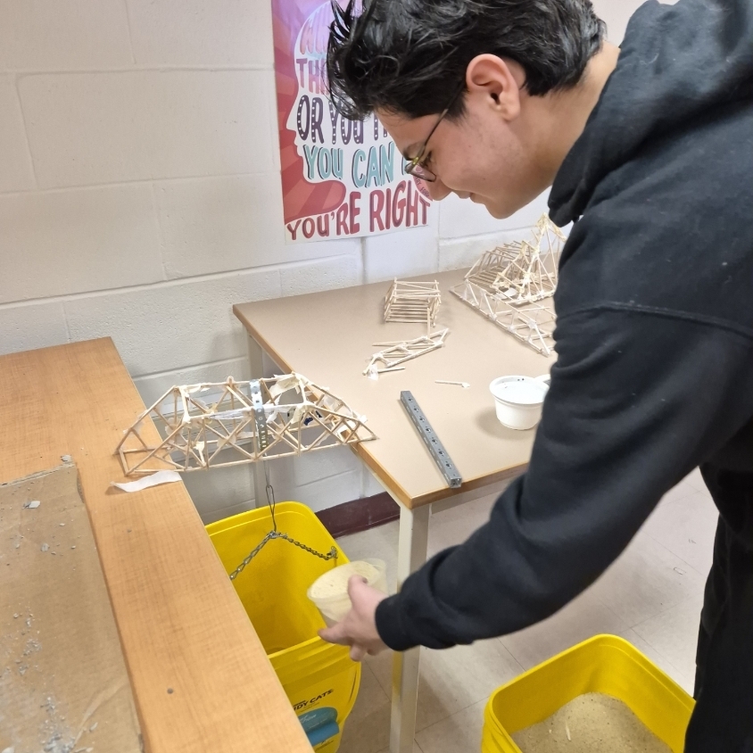 A person wearing glasses and a dark hoodie works on a bridge model in a classroom.