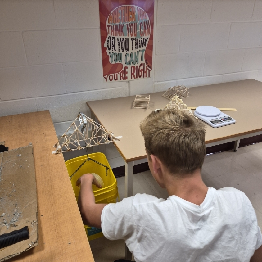 A person leans over a table, building a bridge structure. A yellow bin is nearby.