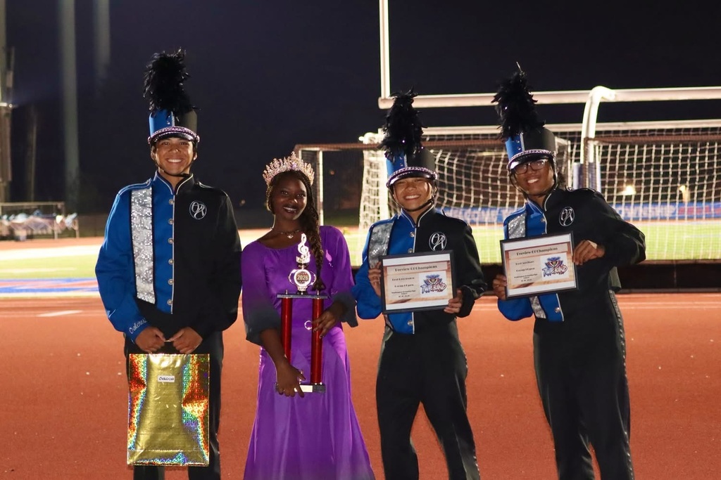 Four people on a field. A woman in a purple dress and three men in black and blue uniforms, holding awards.