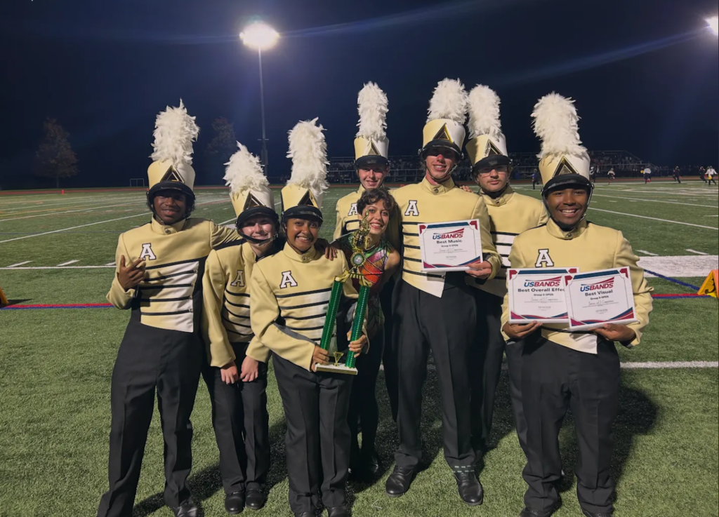 Group of people dressed in marching band uniforms, posing on a field at night, holding certificates and a trophy.