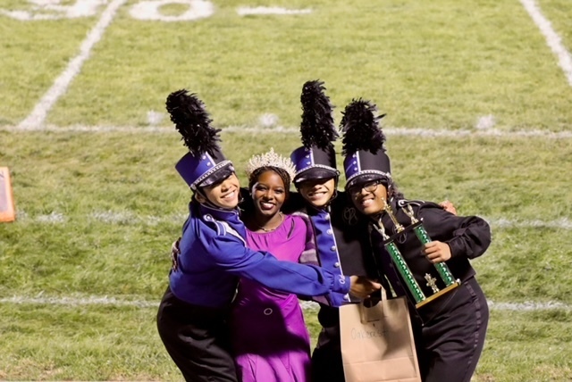 Three individuals in marching band uniforms celebrate on a football field. Two wear hats with feather accents.